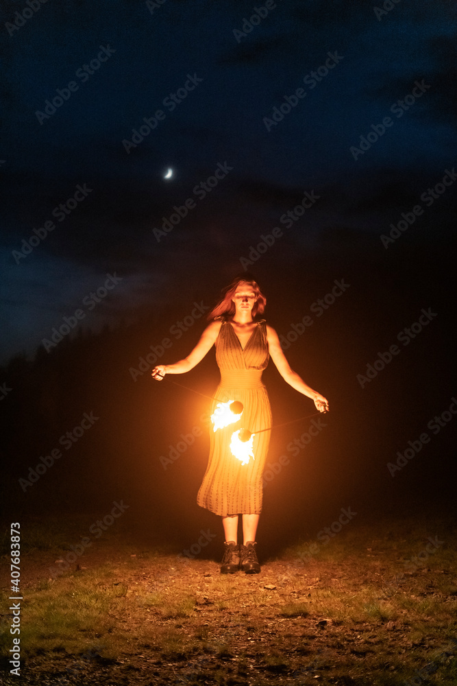 Graceful elegant red haired female fireshow performer making fire dance with two metal fans lit with