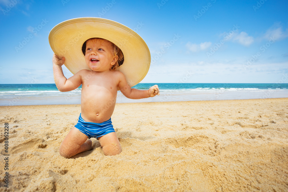 快乐的蹒跚学步的小男孩坐在海滩上，戴着大草帽，背景是大海