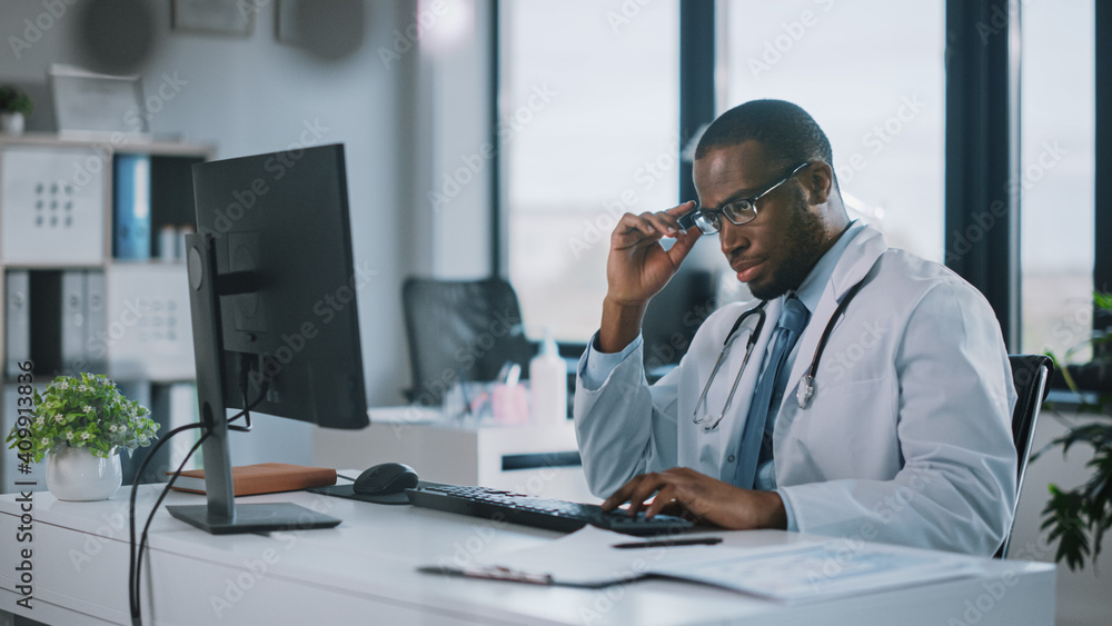 Tired African American Family Medical Doctor in Glasses is Working on a Desktop Computer in a Health