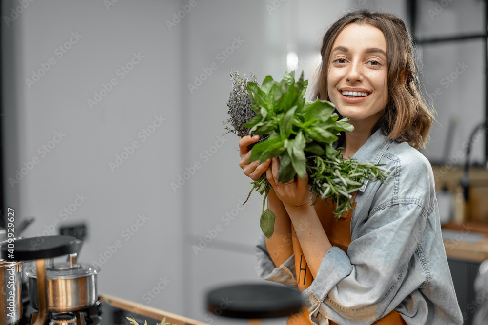 Portrait of cheerful woman in apron with fresh spicy herbs basil, rosemary, thyme on the kitchen. He