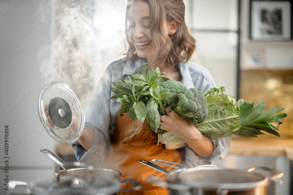 漂亮的家庭主妇穿着围裙，拿着新鲜蔬菜，靠近厨房里有沸腾锅的炊具。健康