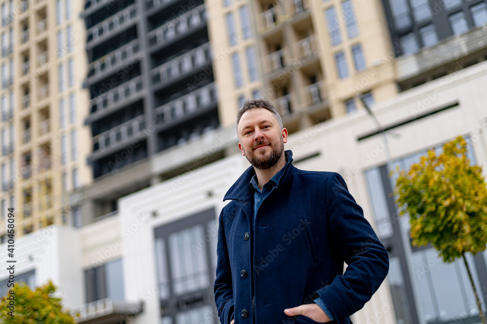 Handsome bearded man in dark blue coat looks to the camera with a smile. Modern building on the back