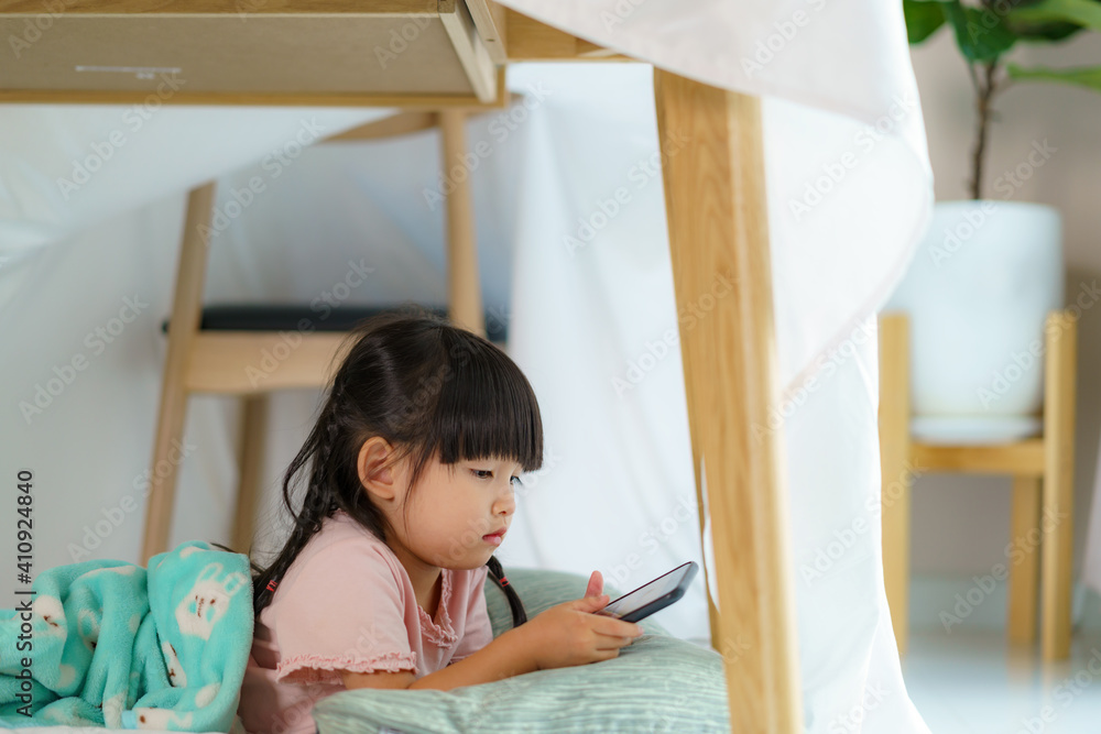亚洲可爱的小女孩躺在客厅a的毯子堡里，用智能手机看动画片
