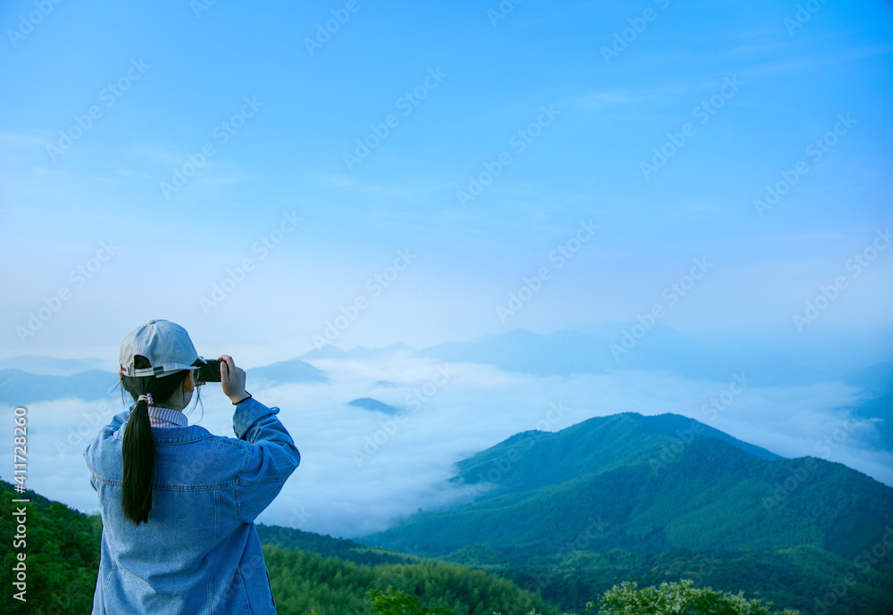 一个戴着帽子的女孩站在山顶，拿着手机拍摄云海和墨海
