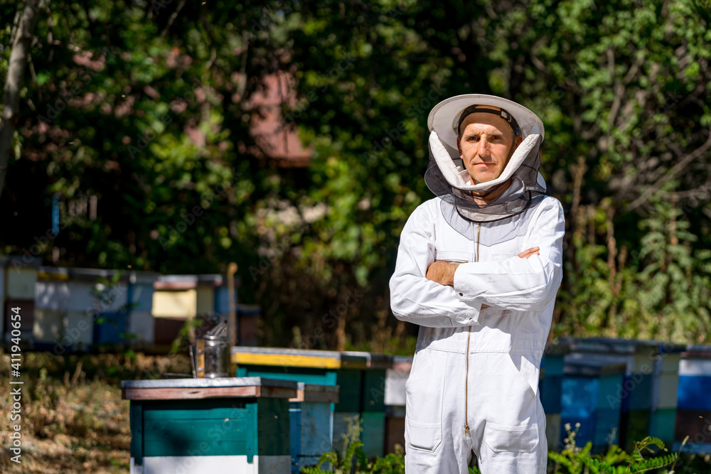 养蜂人站在蜂箱附近的十字线处。雄性身穿白色防护服，周围环绕着绿色t