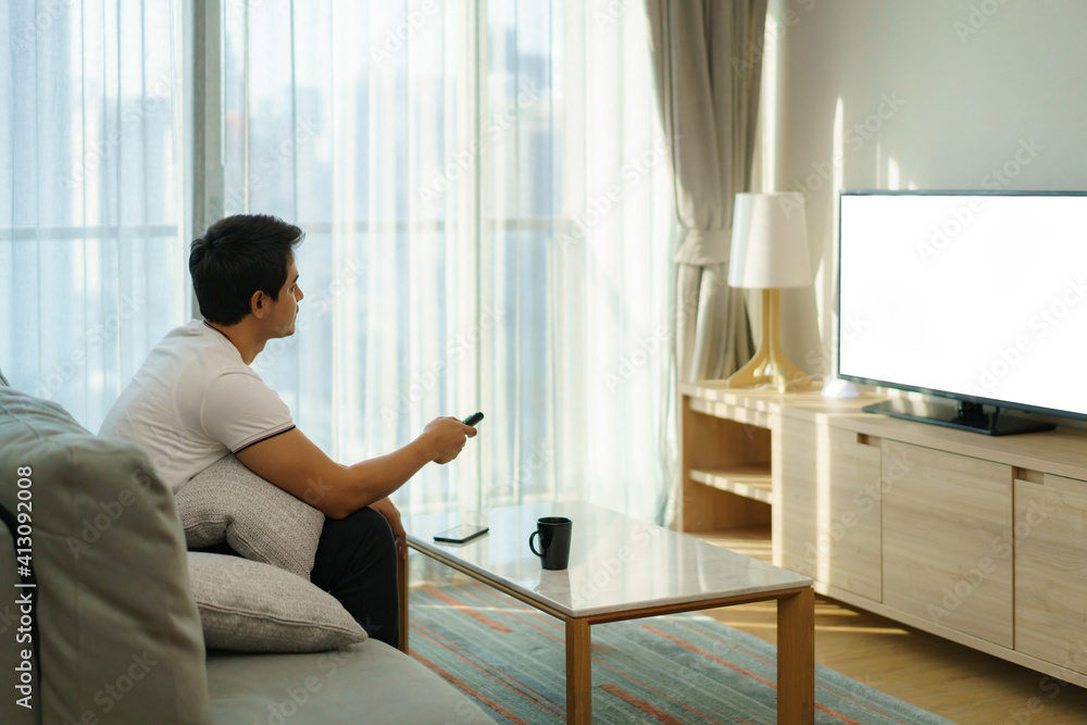 An Asian man holds a TELEVISION remote and is pressing the channel while watching TV on the couch in