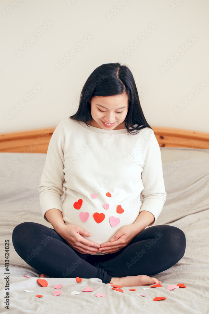 微笑的亚洲中国孕妇坐在床上抚摸自己的肚子。怀孕的肚子上覆盖着