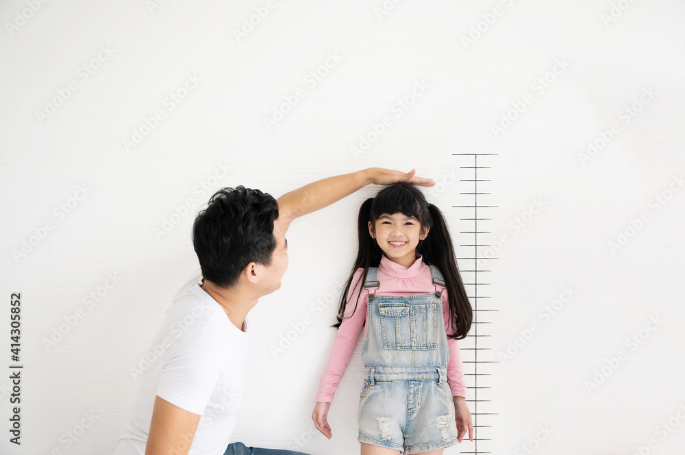 爸爸用秤测量她孩子女儿在白砖墙上的身高增长