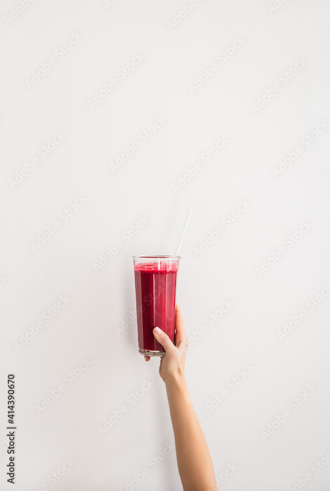 Human hand holding glass with purple beet pomegranate fresh smoothie with glass straw over white wal