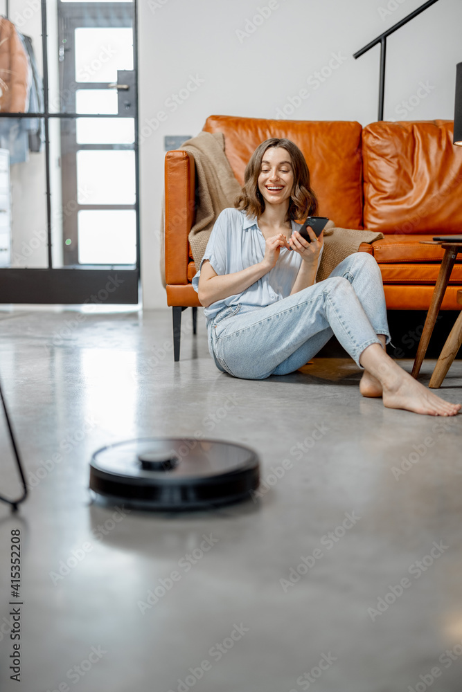 当女人坐在沙发旁使用手机时，黑色机器人吸尘器正在清洁地板。聪明