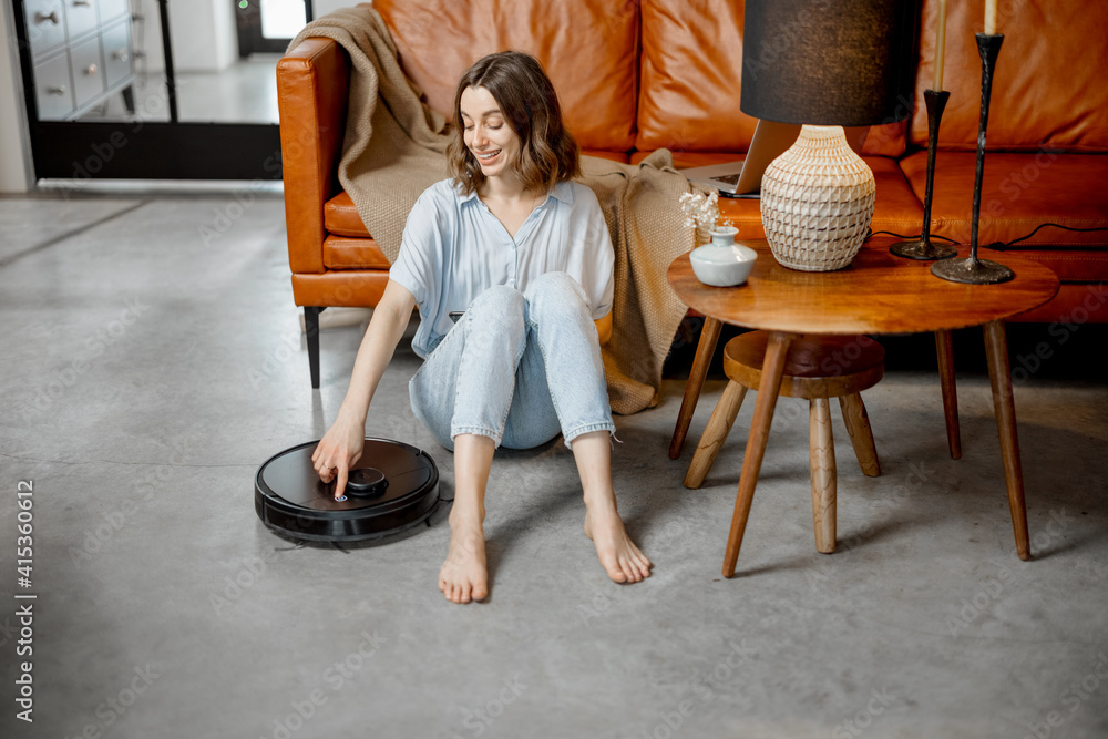 Black robotic vacuum cleaner cleaning the floor while woman sitting near sofa and relax. Smart techn