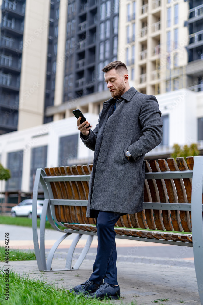 穿着灰色外套、留着胡子的英俊男子靠在长椅上看着手机屏幕。现代建筑o