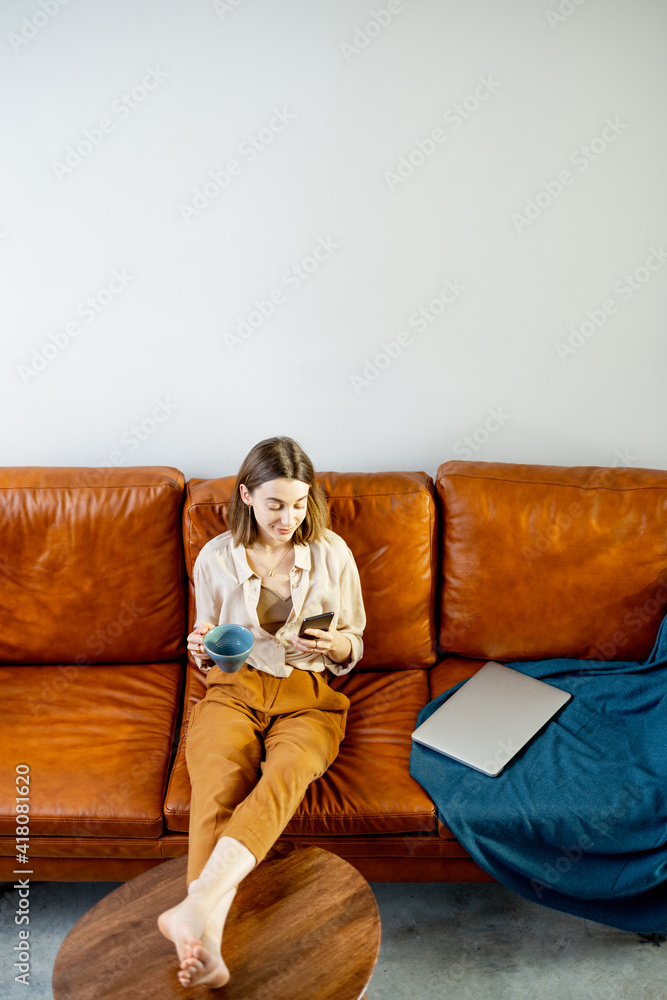 漂亮女人，拿着智能手机坐在家里的棕色沙发上。生活方式和在家工作的概念