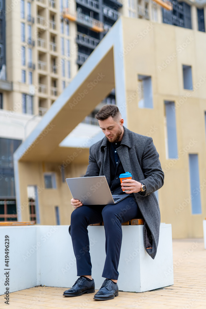 深思熟虑的商人坐在现代化的街道上，拿着笔记本电脑和咖啡。聪明的人在做远程操作