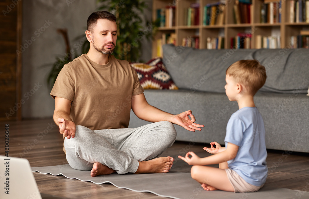 爸爸带着孩子的小儿子在网上做瑜伽，在地板上摆出莲花姿势，闭着眼睛冥想