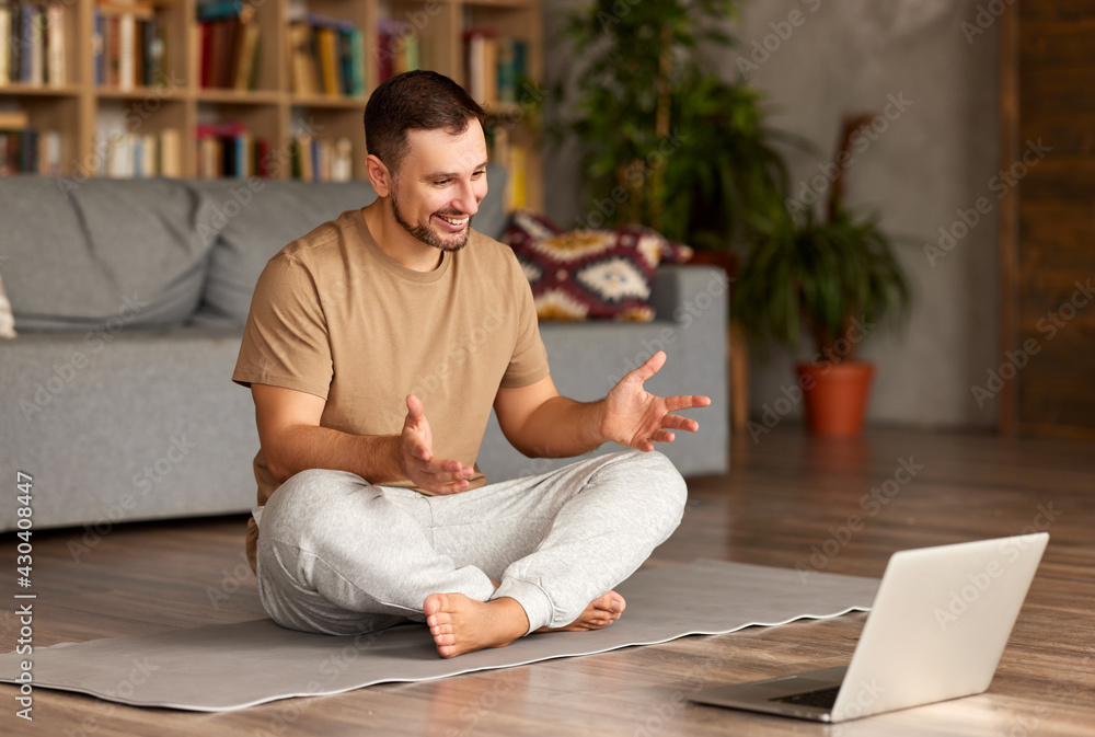 一名穿着休闲服的年轻男子以莲花姿势坐在笔记本电脑前，在线练习瑜伽