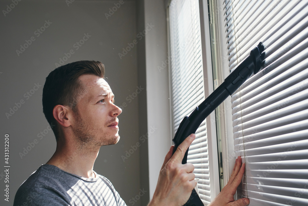 男人在家用吸尘器清理百叶窗上的灰尘。主题是家务和家政。