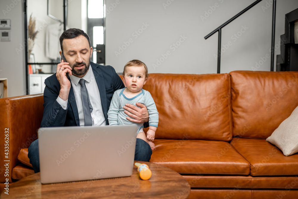 Young dad working on a laptop and speaking on phone at home while taking care of his baby son. Multi