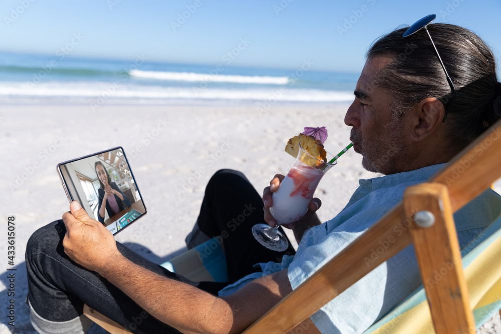 Caucasian man relaxing on beach with drink having video call using tablet