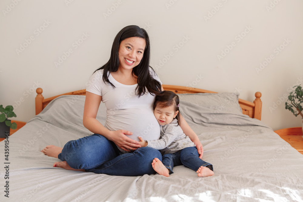 亚洲华裔孕妇，家里有一个蹒跚学步的女孩坐在床上。女孩女儿孩子拥抱妈妈