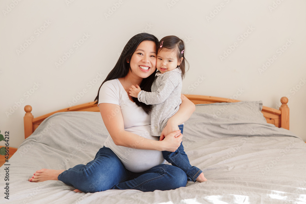 亚洲华裔孕妇和蹒跚学步的女孩在家里的床上拥抱。女孩女儿孩子在玩机智