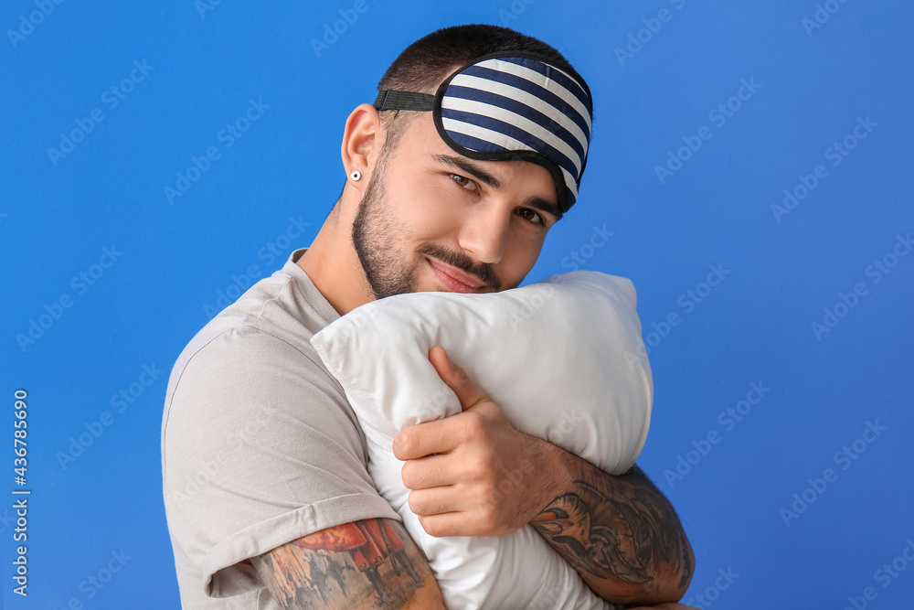 Young man in sleeping mask hugging pillow on color background