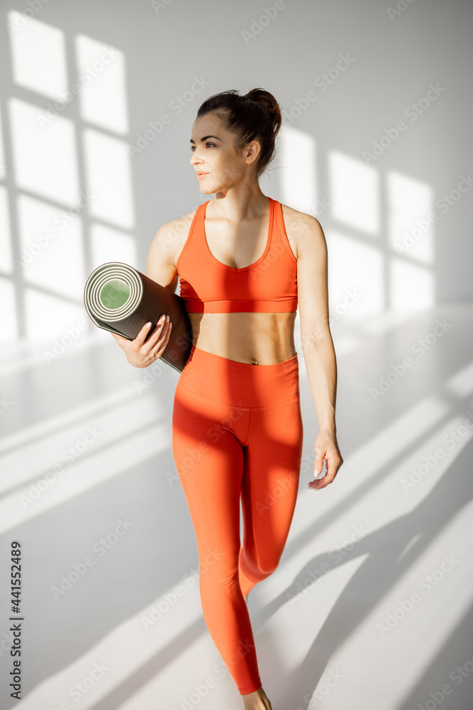 一位身穿明亮运动服的运动女性的生活方式肖像，在白色健身房里拿着瑜伽垫行走