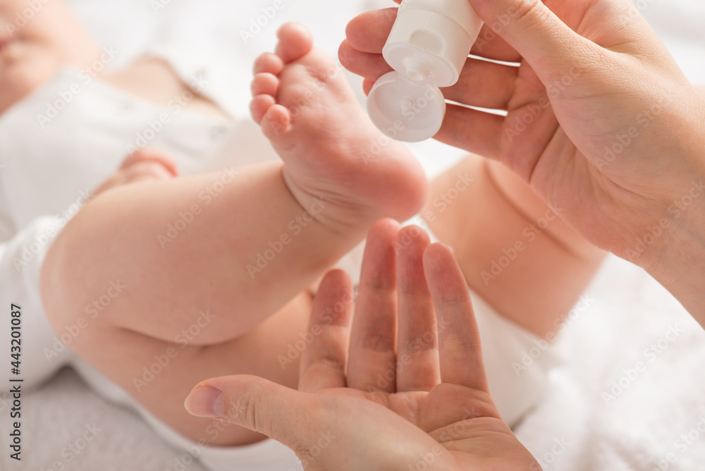新生儿的腿、脚和母亲的手在隔离的wh上拿着白色奶油管的特写照片