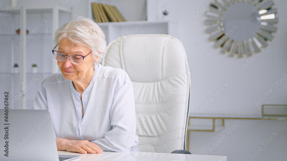疲惫困倦的老太太经理在白色桌子上的灰色笔记本电脑上打字，坐在大椅子上对着dif