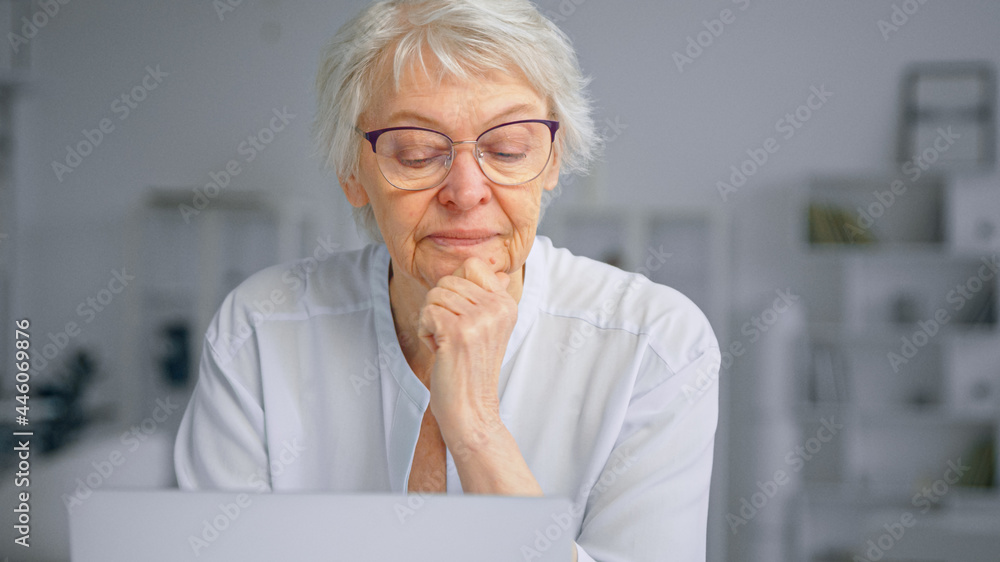 一位留着灰色短发的老女商人头枕着手看着窗户，坐在笔记本电脑旁