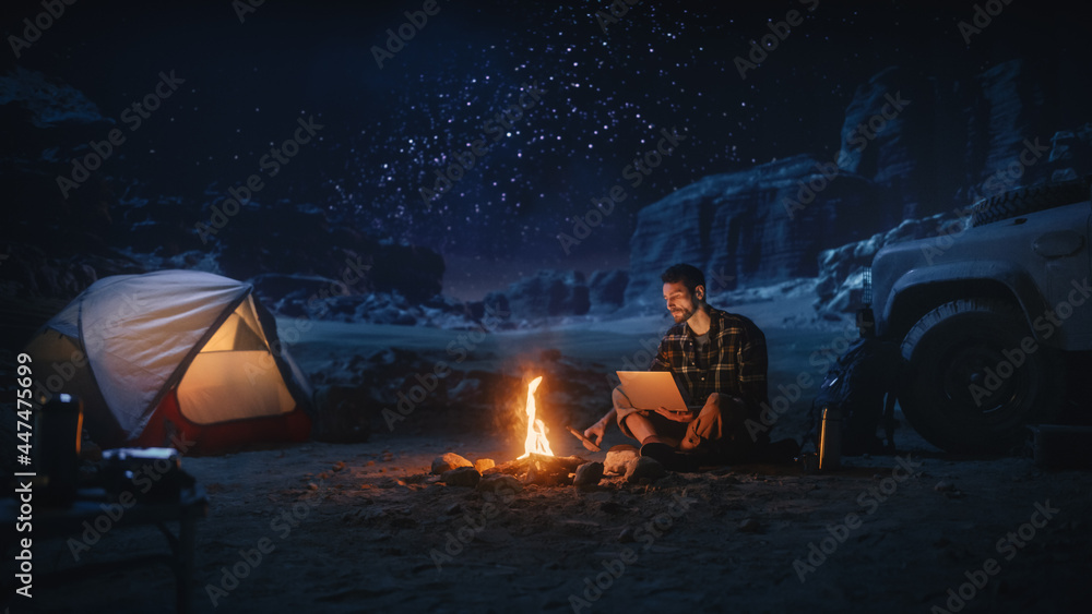 坐在篝火旁的男性旅行者在峡谷露营时使用笔记本电脑。男人在做D