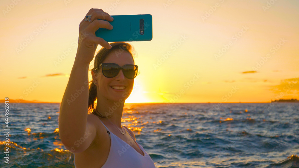 特写：日落时，微笑的年轻女子在海边自拍。