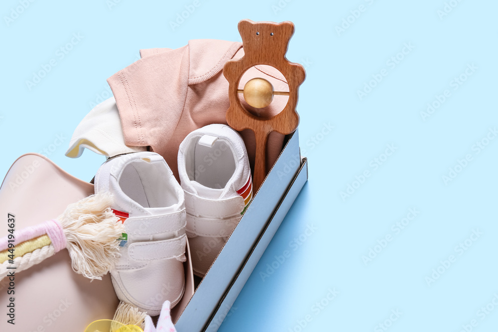 Box with baby clothes, shoes and toy on color background, closeup