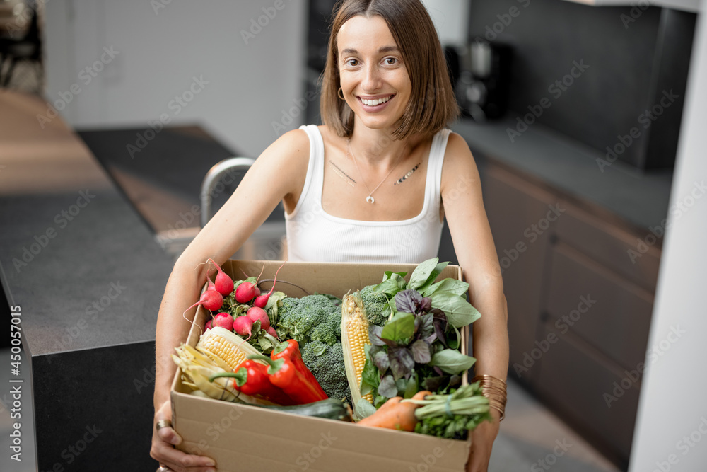 Portrait of a young woman with fresh vegetables packed in cardboard box at kitchen. Delivering and b