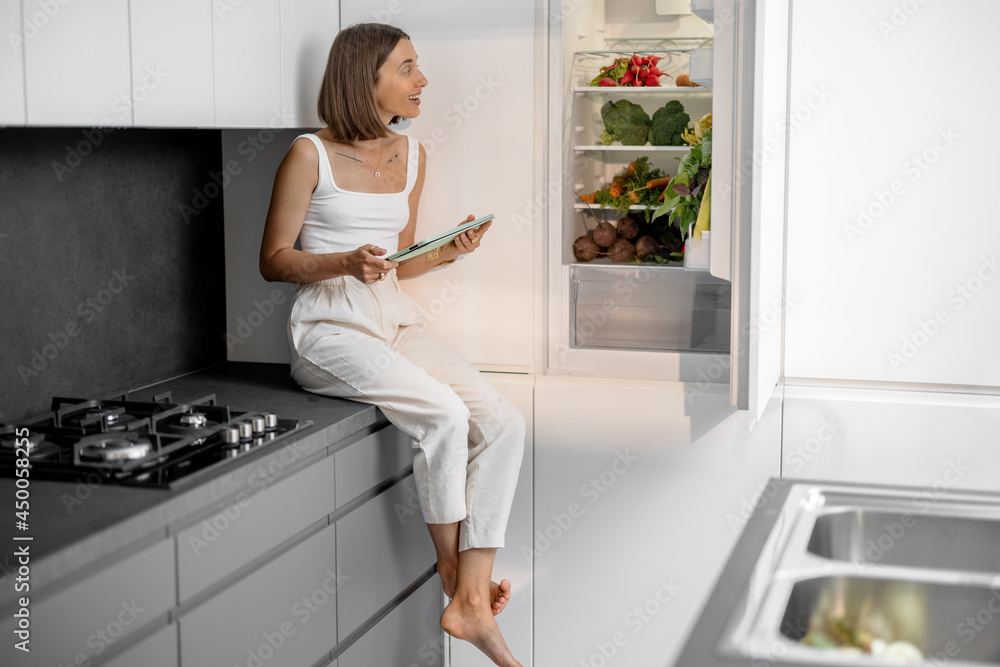 Young woman sitting near the fridge full of fresh vegetables at modern kitchen. Healthy vegan eating