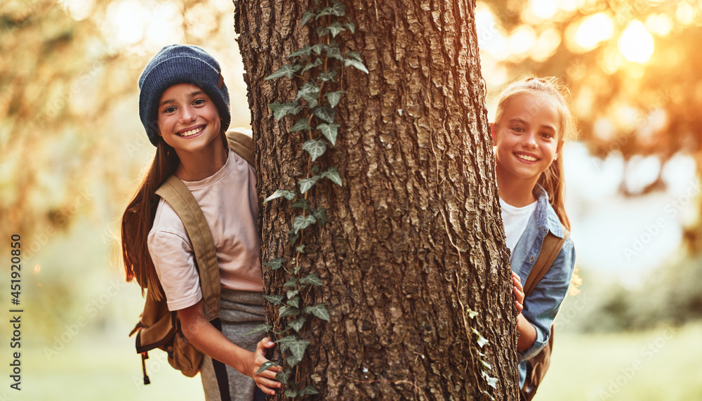 Happy school girls hugging tree in forest and smiling at camera during camping activity in nature