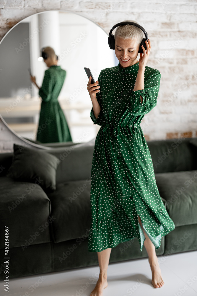 穿着绿色连衣裙的时尚女性戴着耳机欣赏音乐，在客厅里用手机跳舞