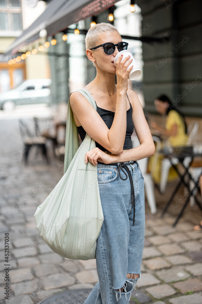 时尚时尚的女人带着咖啡和包包在户外散步。街头时尚和生活方式概念