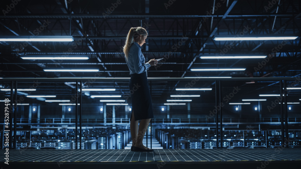Female IT Specialist Using Tablet Computer in Data Center, Walking on a Bridge Overlooking Gigantic 