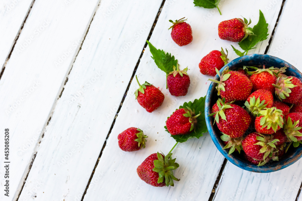 成熟的红色草莓放在白色木制桌子上的蓝色杯子里。附近散落着甜浆果。