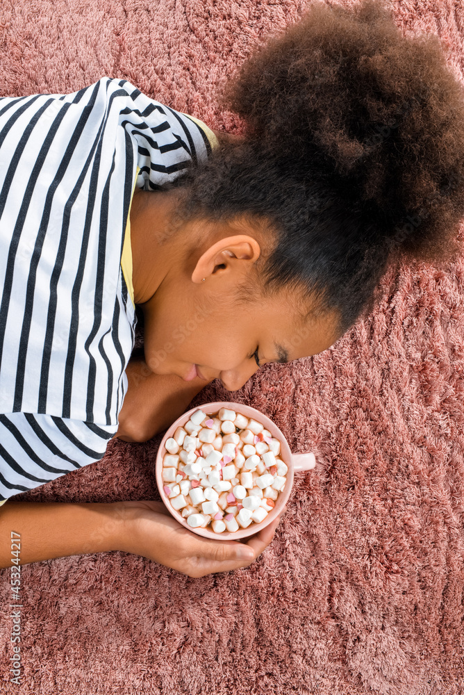 非洲裔美国少女躺在地毯上，手里拿着一杯热可可，俯视图