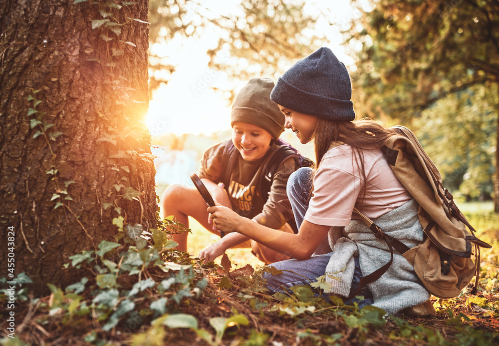 两个戴着保暖帽子、背着背包的小女孩在森林里用放大镜检查树皮