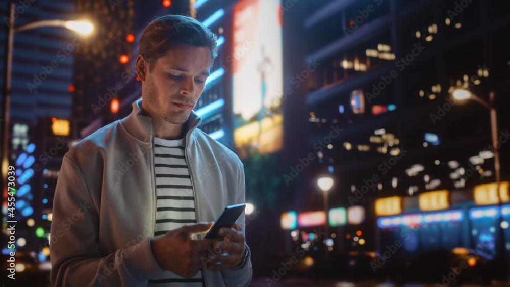 使用智能手机的男人在充满霓虹灯的夜城街道上行走。微笑时尚的男人在使用