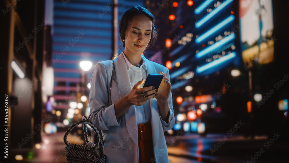 年轻漂亮的女人用智能手机穿过充满霓虹灯的夜城街道。微笑