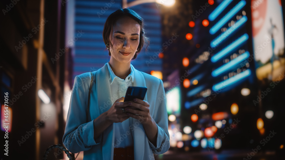 年轻漂亮的女人用智能手机穿过充满霓虹灯的夜城街道。微笑