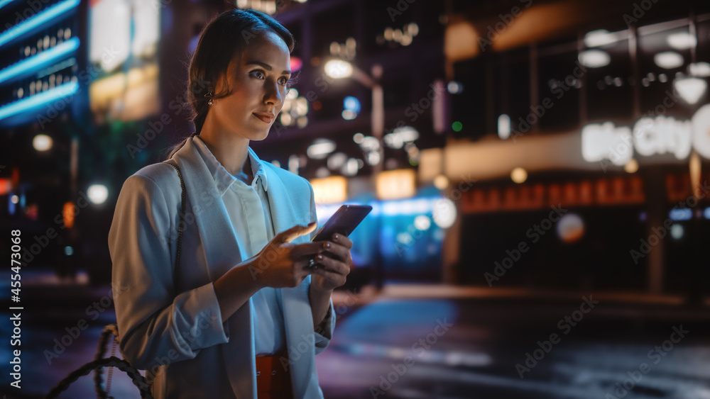 漂亮的年轻女人用智能手机穿过充满霓虹灯的夜城街道。波特拉伊