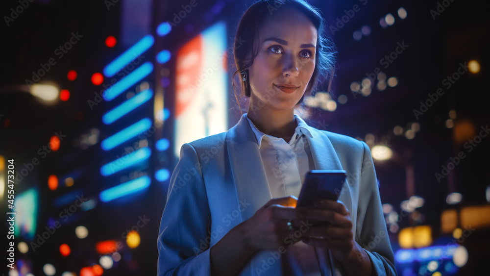 美丽的年轻女子用智能手机站在霓虹灯照亮的夜城街道上。波特拉伊