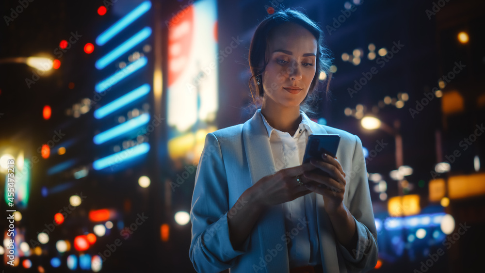 美丽的年轻女子使用智能手机站在充满霓虹灯的夜城街道上。波特拉伊