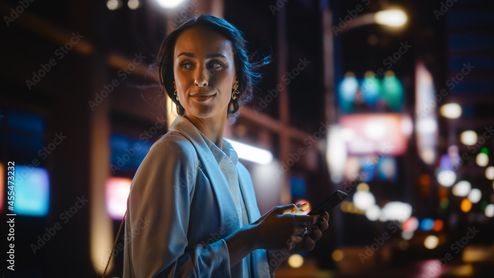 美丽的年轻女子用智能手机穿过充满霓虹灯的夜城街道。微笑
