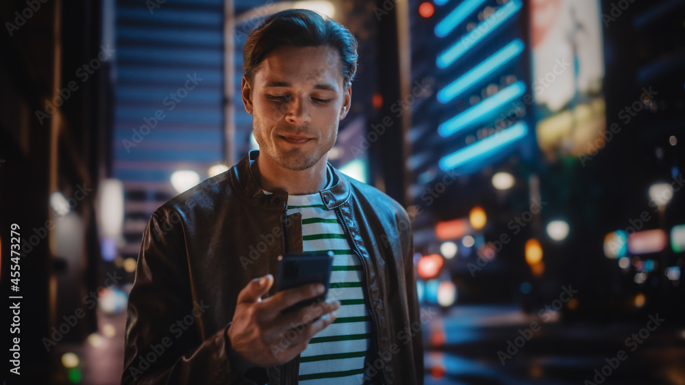 使用智能手机的男人在充满霓虹灯的夜城街道上行走。微笑时尚的男人在使用